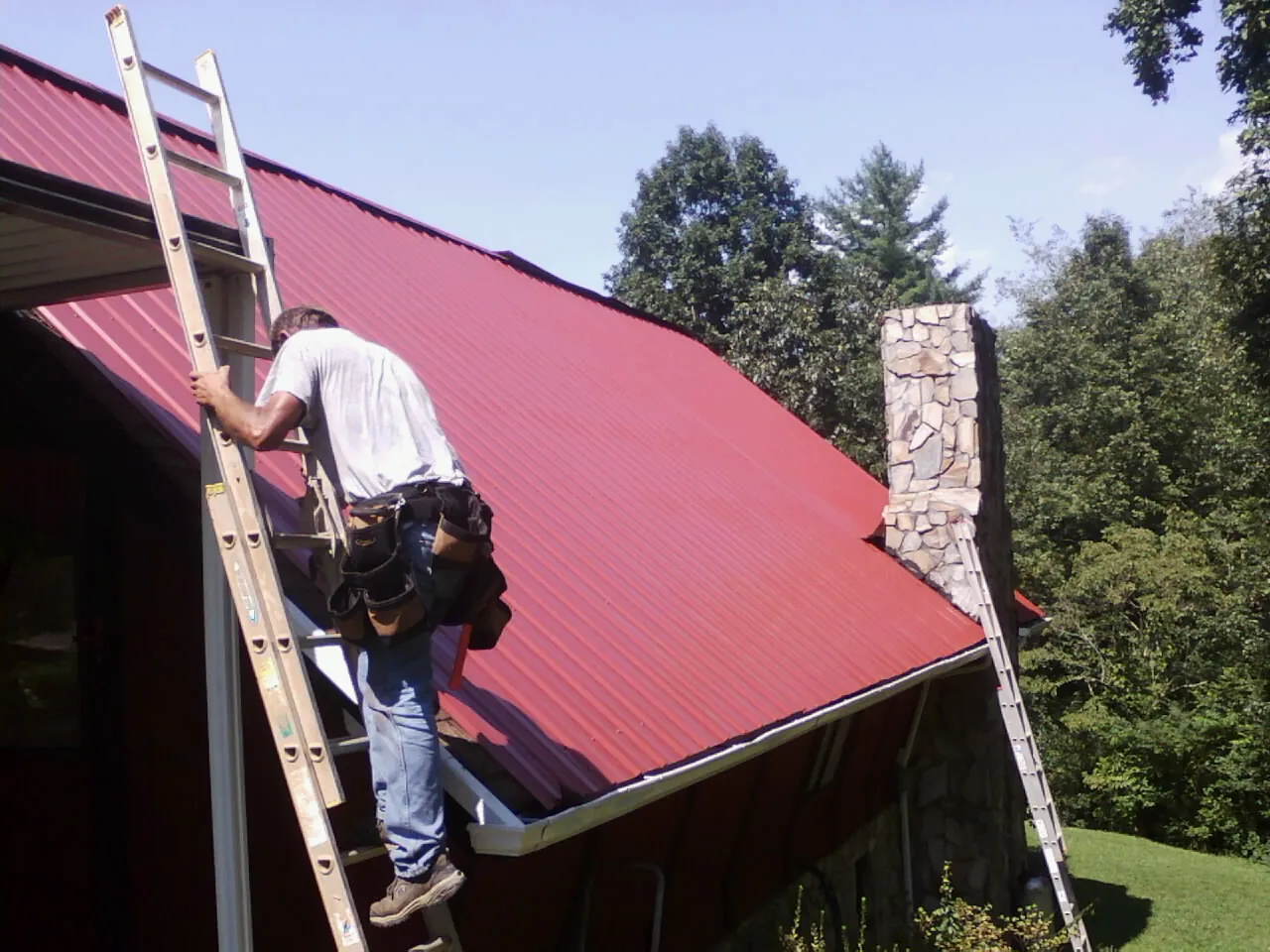 Roofer on a ladder beside a red metal roof and stone chimney — Soffit & Fascia Repair by Roofing Brothers in Latta